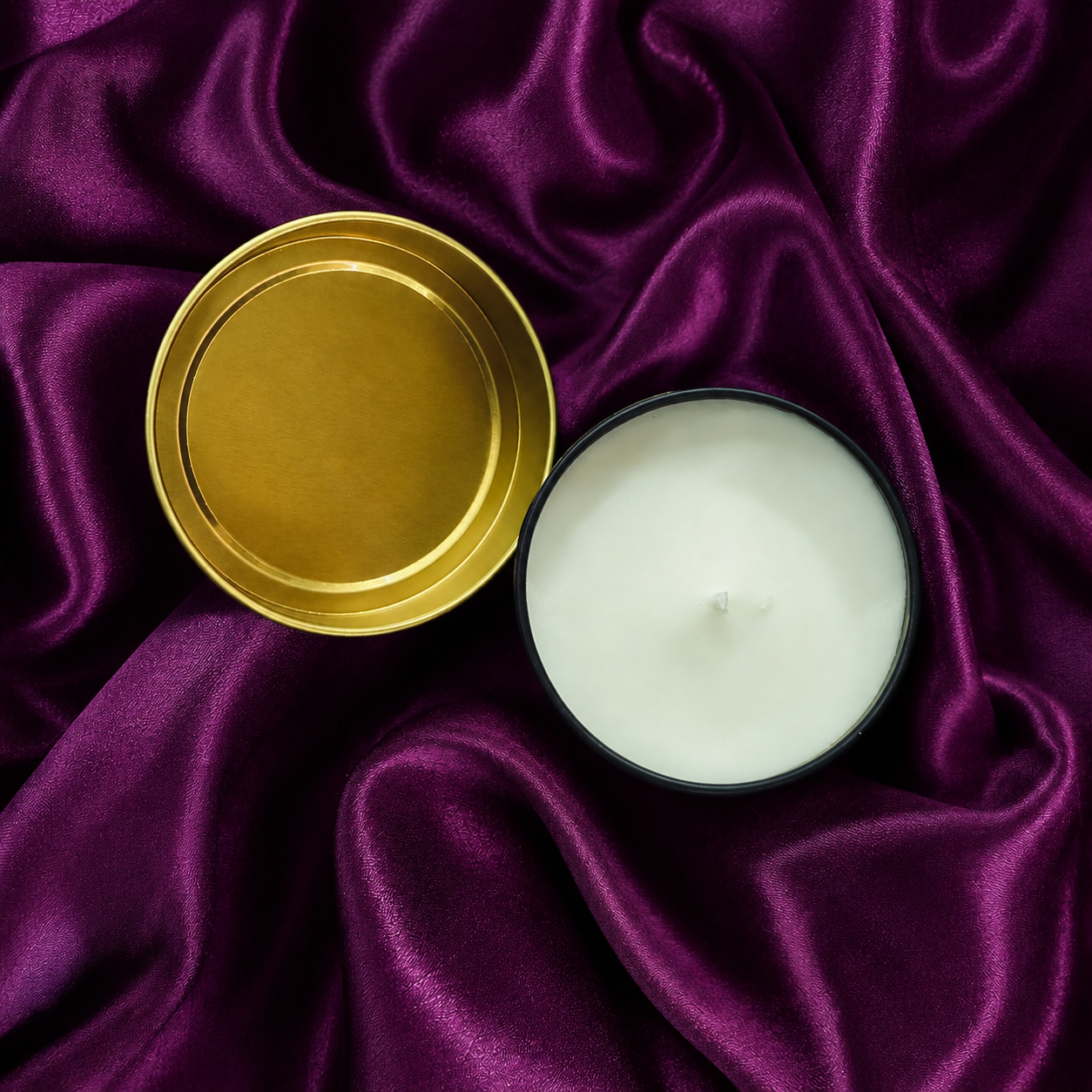 An open candle tin with white wax beside its gold-colored lid, set on a draped purple satin fabric background.