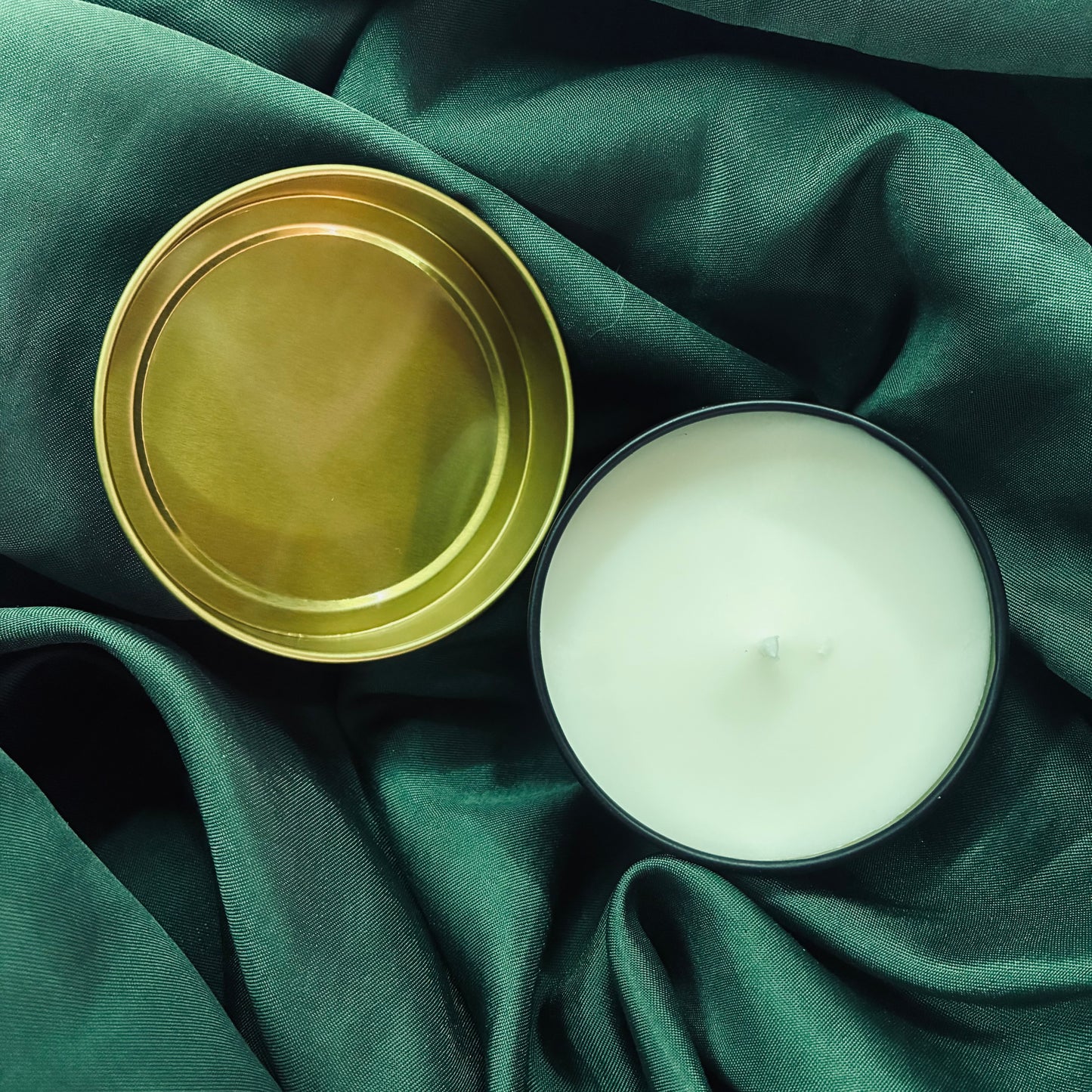 An open black tin candle with white wax sits beside its gold-colored metal lid on a draped dark green fabric background.