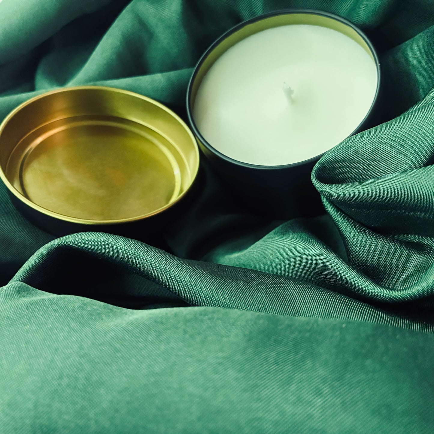 A white wax candle in a dark metal tin next to its gold-toned lid, styled on draped green fabric.
